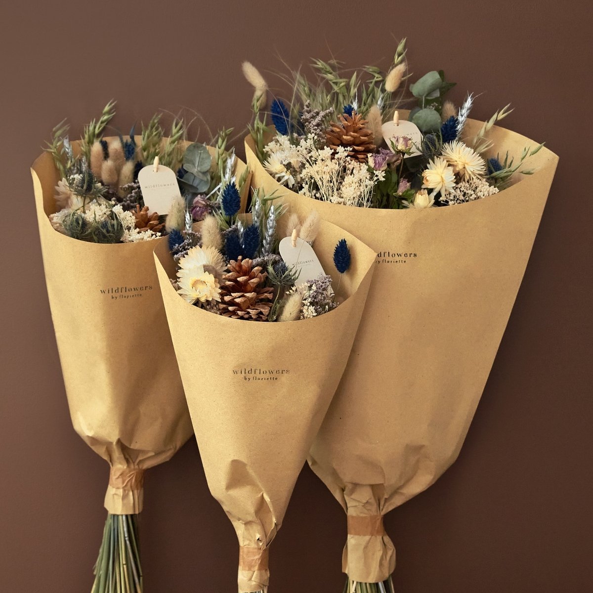 Three holiday field bouquets with pinecones, blue phalaris, and strawflowers in kraft paper wraps